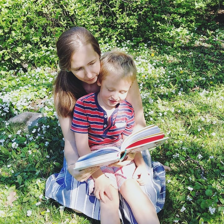mom in a dress sitting on a patch of clover with a little boy in a striped tshirt on her lap, reading a hardcover book together