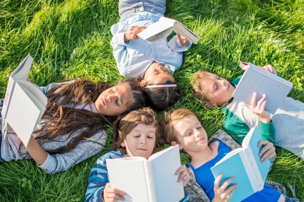 5 kids lying on green grass reading hardcover books in a circle